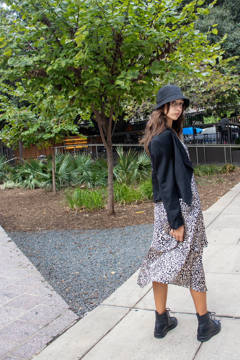Woman in a black jacket and patterned skirt standing on a sidewalk with trees in the background in a black bucket hat